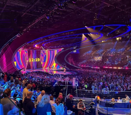Emma and Lizzie's view from M & S Bank Arena wheelchair seating section 5 for Eurovision. The entire stage is visible and lit in a bright pattern. There is a screen at the back of the stage counting down to the start of the show. The crowd fills the entire arena.