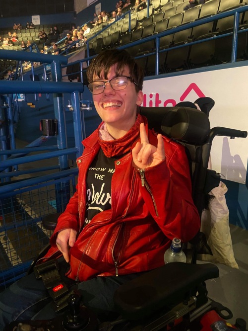 Emma, a young woman with brunette hair and glasses, is sat in her wheelchair at the Utilita Arena Birmingham smiling broadly and throwing the "metal horns" hand signal at the camera. She is wearing a red leather jacket, black jeans, black Bring Me The Horizon band t shirt and a red face covering around her neck.