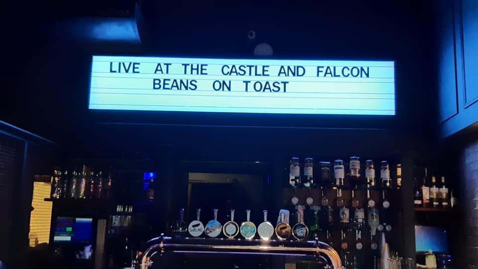The bar in the concert room at Castle and Falcon. Above the bar is a sign (black text on a white background) which reads " LIVE AT THE CASTLE AND FALCON BEANS ON TOAST"