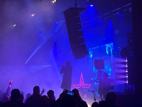 Photo from Starset at KK's Steel Mill on 2nd March 2023. Side-angle view of the stage, backlit by blue and purple light. The lead singer is silhouetted in profile, holding a mic with a Star logo outlined in pink light behind him. There are speakers and lighting rigs hanging from the ceiling and the side of the stage. Fans are silhouetted in front of the stage.