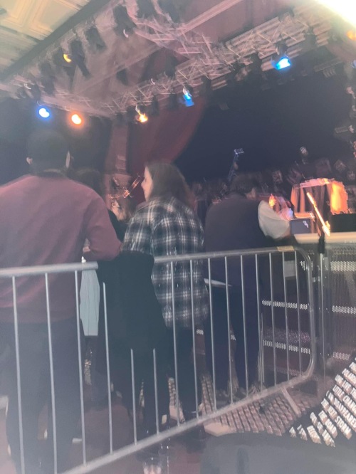 Emma's view from the access area at Ipswich Corn Exchange. There is a large crowd beyond the barrier in front of Emma. Microphones are just visible on stage above the crowd