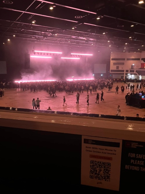 Emma's view from accessible seating at the Utilita Arena Cardiff. The floor standing section is visible below the balcony, with a crowd of fans in front of the stage. The stage itself is light in a pink/purple hue and looks cloudy as though smoke machines have just been tested