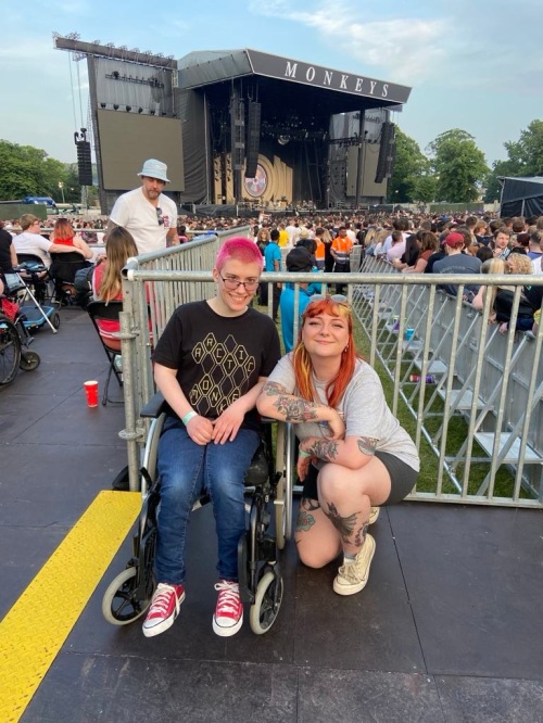 Emma and Dev on the access platform at Arctic Monkeys Hillsborough Park, with the stage behind them. Emma is sat in her manual wheelchair and Dev is kneeling beside her.